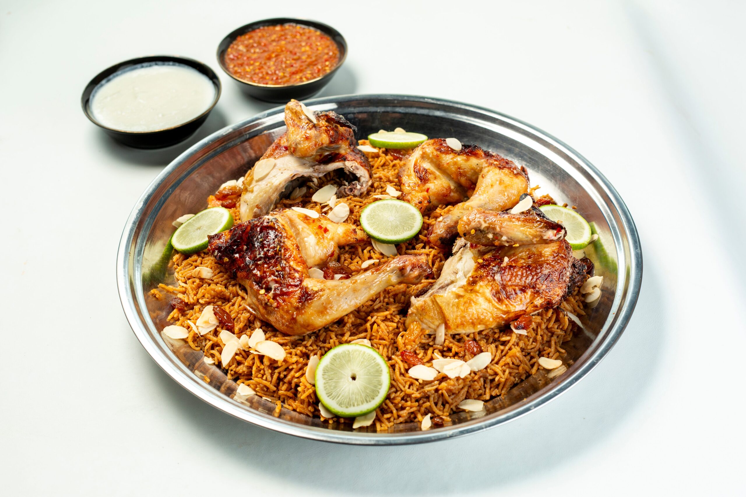 Close-up of chicken biryani with slices of lime, almonds, and sauces on a white background.